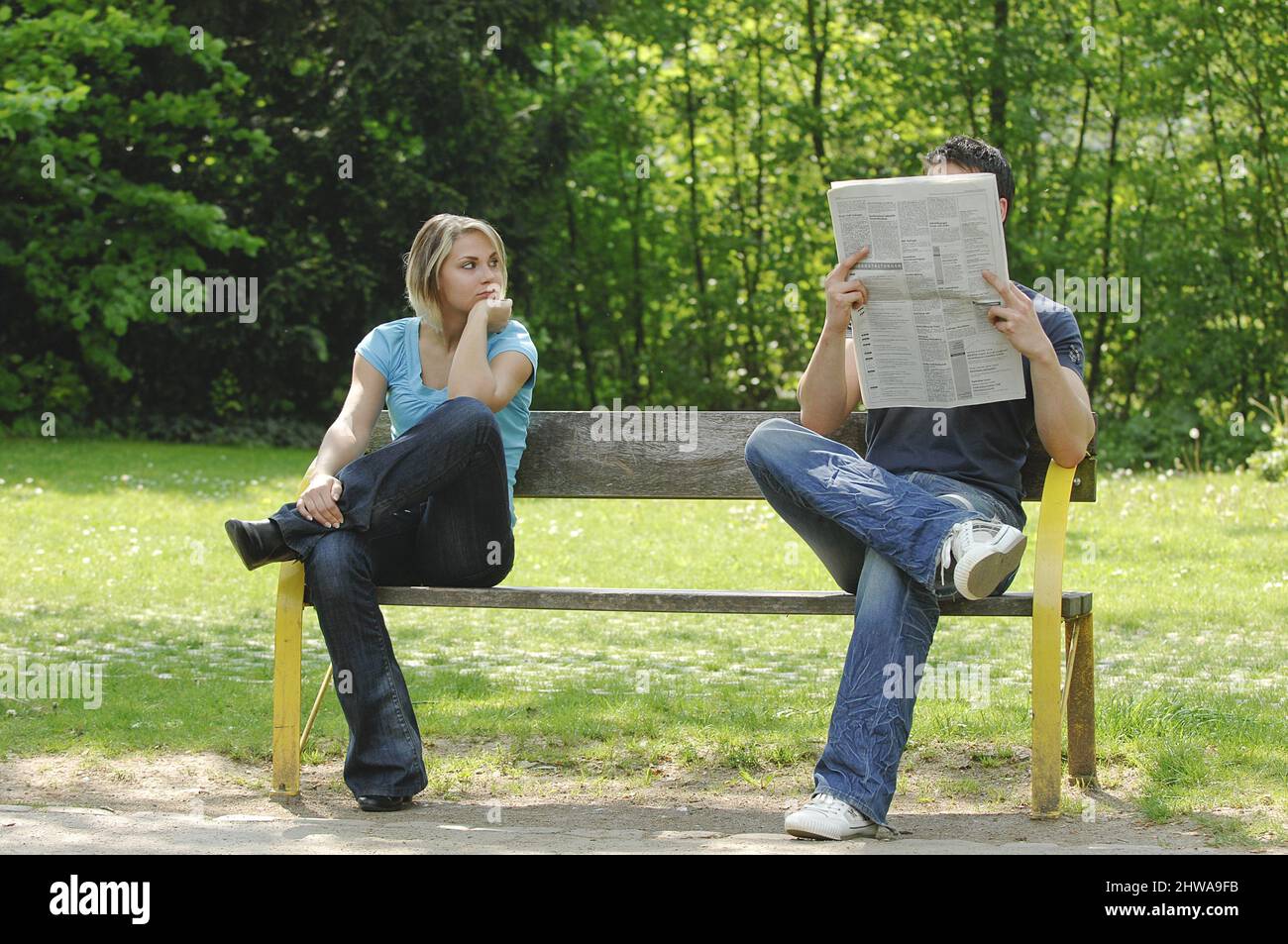 quarrelling couple sitting far apart on a park bench, he reads the newspaper, she looks at him expectantly Stock Photo