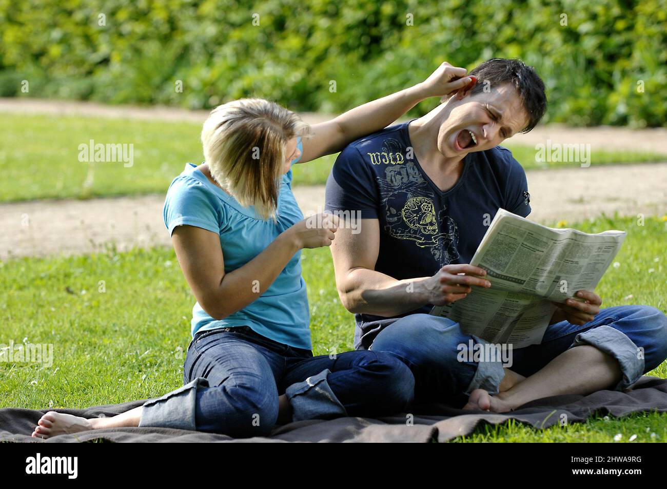 couple sitting on a meadow, he reading the newspaper, she pulling his ears out Stock Photo