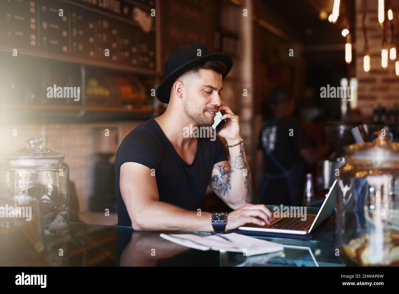 Busy behind the bar. Shot of a young man using a phone and laptop while working in a coffee shop. Stock Photo