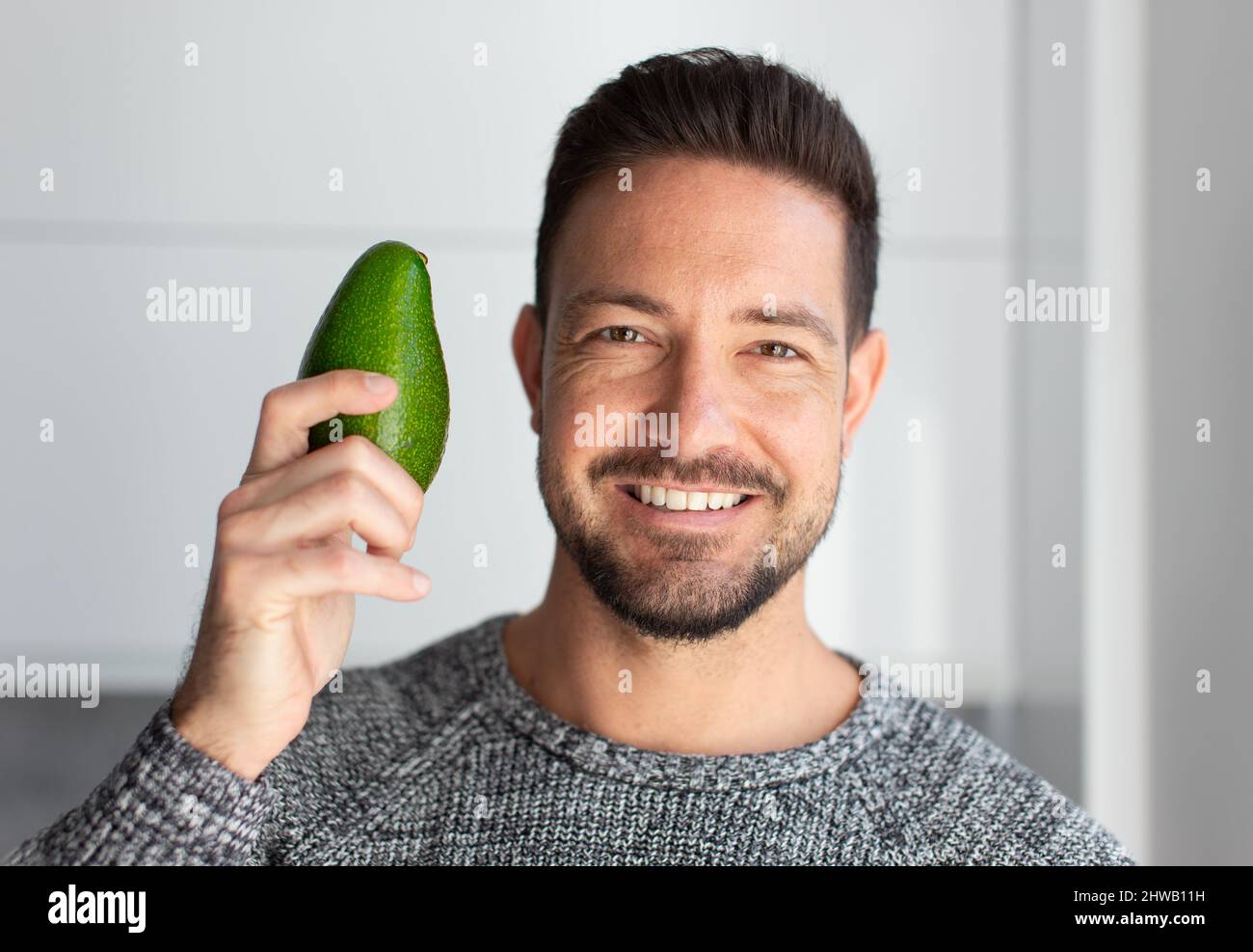 Happy young man with toothy smile holding avocado in kitchen portrait Stock Photo