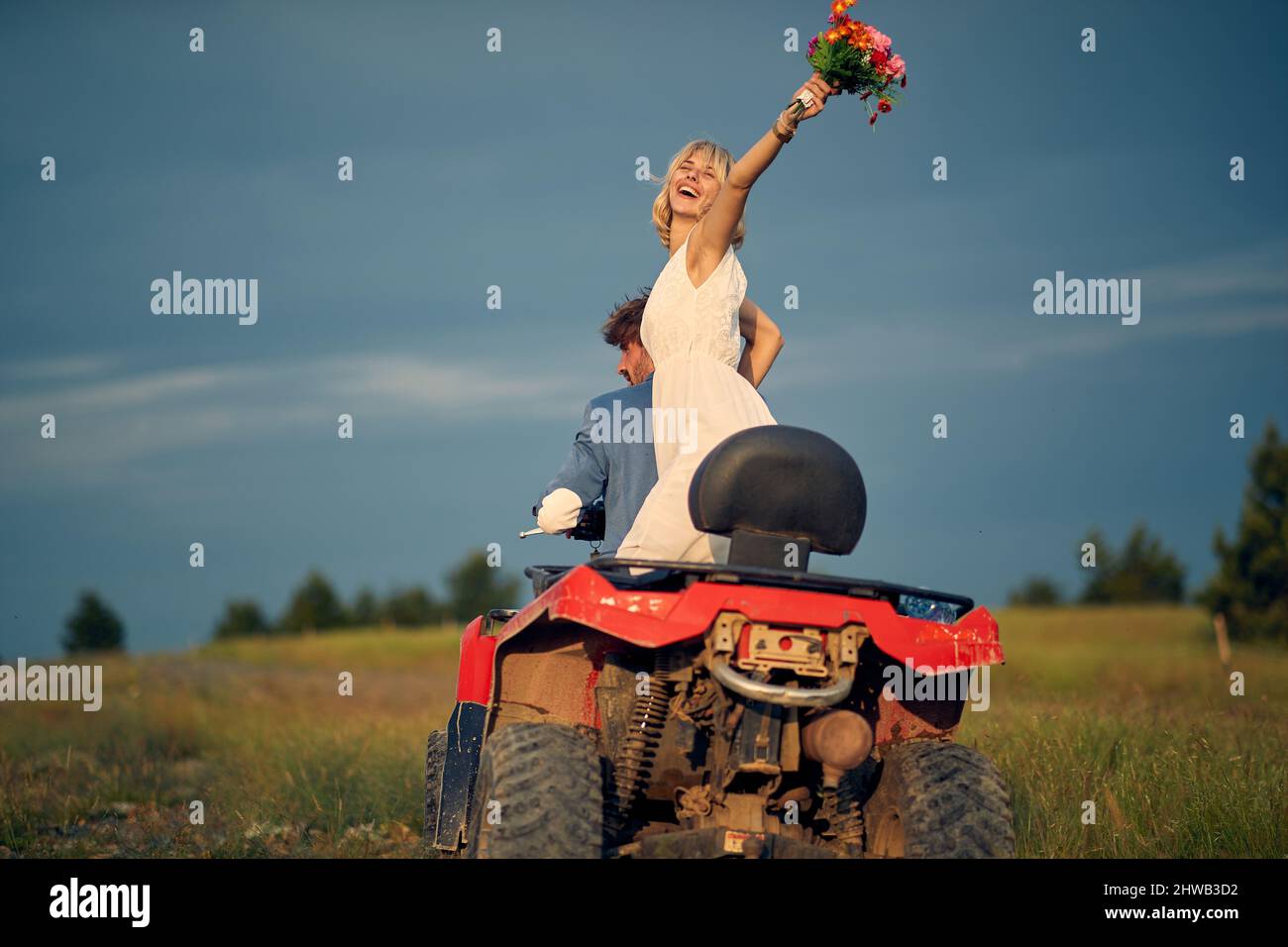 Happy bride throwing bouquet into air. Joyful newlywed couple riding quad. Love, marriage, wedding, happiness, couple concept. Stock Photo