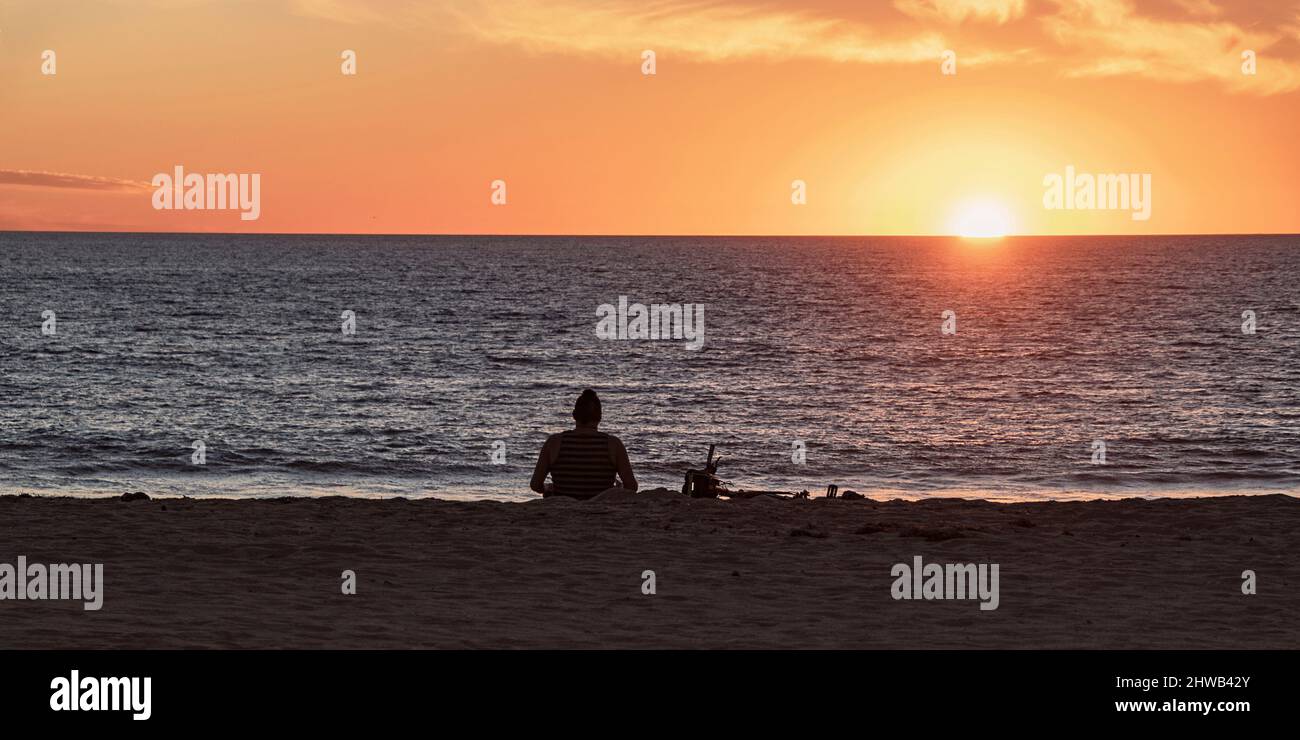 silhouette of a man sitting alone on a Southern California beach watching a blazing orange sunset with a dark sandy foreground Stock Photo