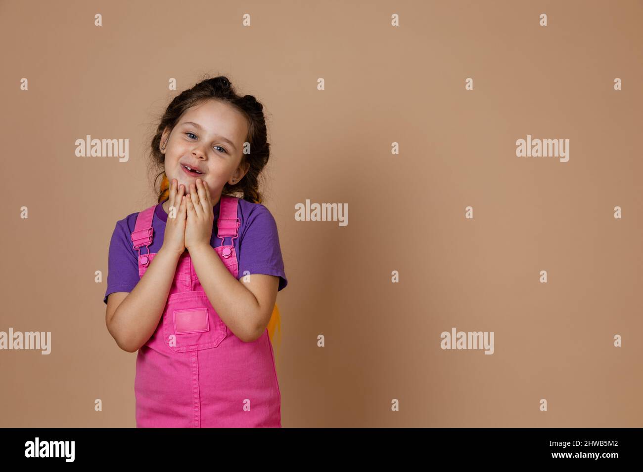 Kind cute, small girl with kanekalon pigtails of yellow color, touching chin with hands looking at camera with gentle smile wearing pink jumpsuit and Stock Photo