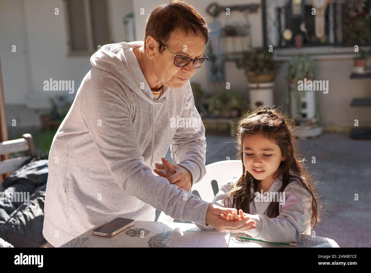 Beautiful girl writing and counting on fingers with the help of her grandmother on an outdoor pergola. Senior teacher supervising little kid doing homework back yard. Happy micro moments of life. Stock Photo