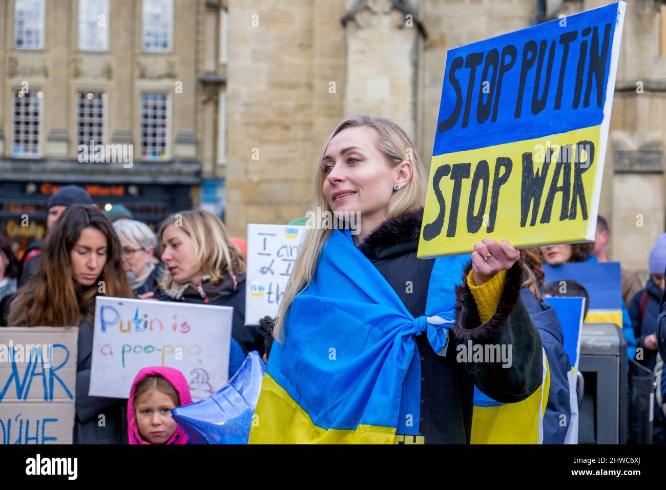 Bath, UK. 5th March, 2022. A woman draped in the Ukrainian flag holds an anti-Putin / stop the war placard as she listens to speeches in front of Bath Abbey during a demonstration against Russia's invasion of Ukraine. The demonstration was organised in order to allow local people to show their solidarity with Ukraine in the Russia-Ukraine war and to condemn Putin's actions. Credit: Lynchpics/Alamy Live News Stock Photo