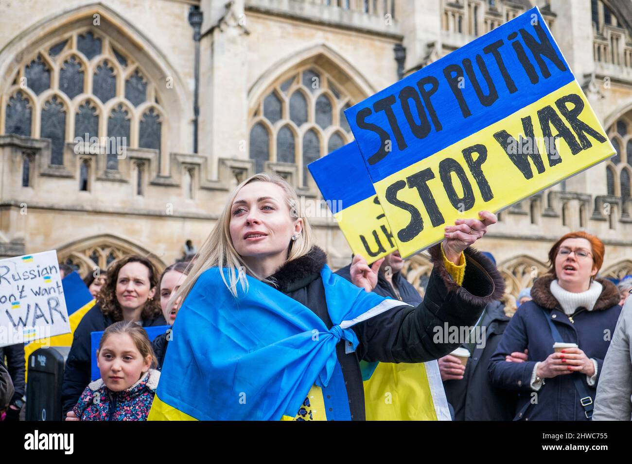 Bath, UK. 5th March, 2022. A woman draped in the Ukrainian flag holds an anti-Putin / stop the war placard as she listens to speeches in front of Bath Abbey during a demonstration against Russia's invasion of Ukraine. The demonstration was organised in order to allow local people to show their solidarity with Ukraine in the Russia-Ukraine war and to condemn Putin's actions. Credit: Lynchpics/Alamy Live News Stock Photo