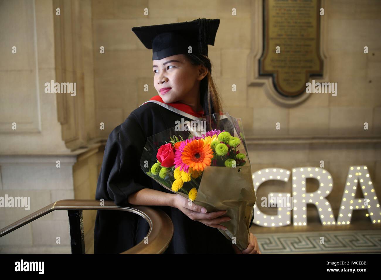 A pretty Asian female graduate holding a bouquet of flowers poses for