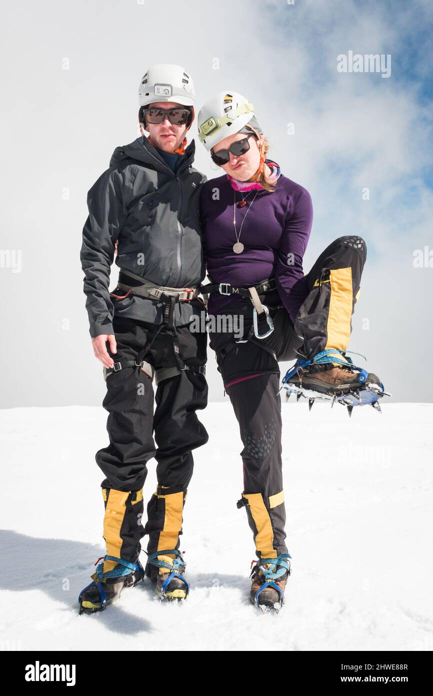 Couple in hiking gear proudly and energetically show crampons on snowy summit Stock Photo