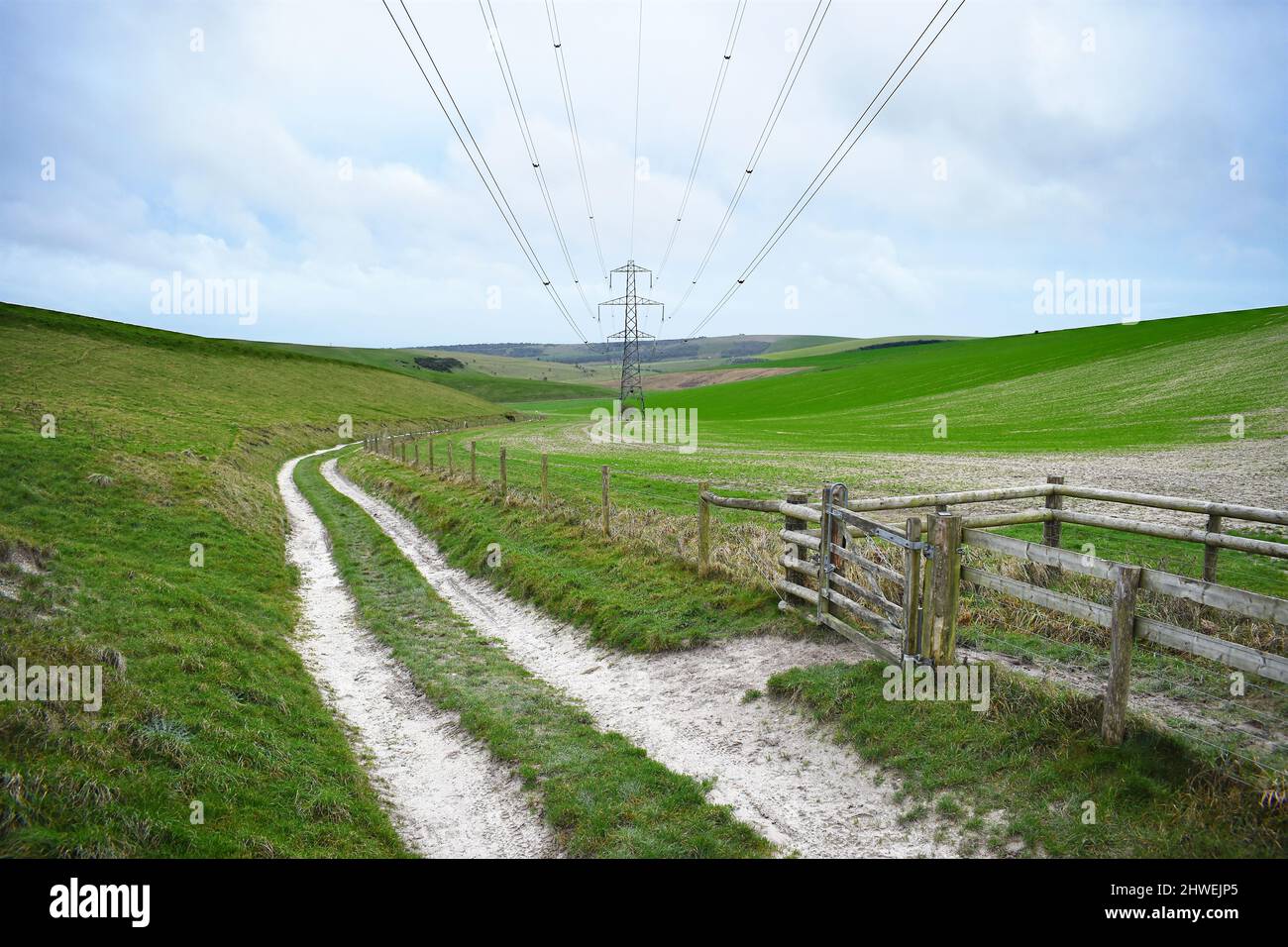A rutted track next to a footpath gate runs into the distances underneath electric cables in the sky with a electricity pylon in the background. Stock Photo