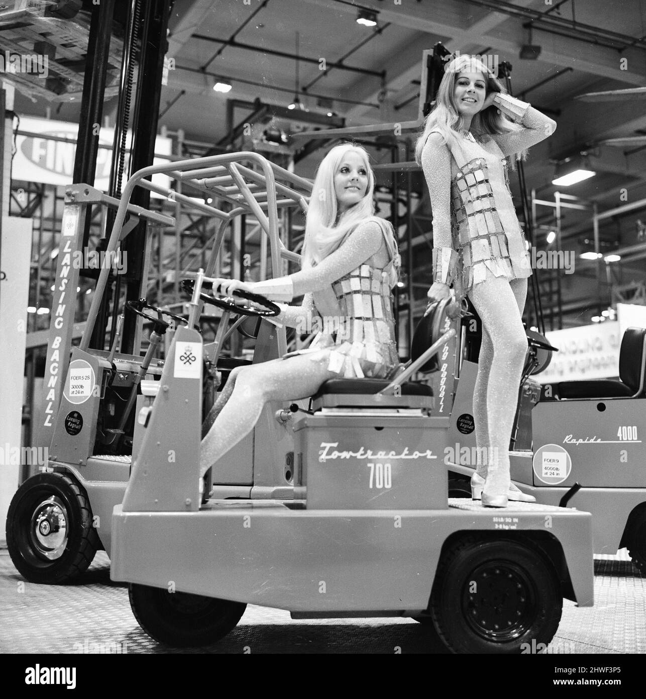 Mechanical Handling Exhibition, Earls Court, London, Tuesday 5th May 1970. Our Picture Shows ... Valerie Pugh giving a lift to Jenny Mould on an electric tow tractor on the Lancing Bagnall stand at the exhibition, the models are wearing suits on silver lurex and plastic. Stock Photo