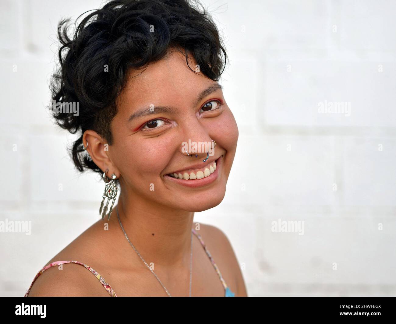 Young positive optimistic beautiful fresh Mexican Latina woman with short curly hair and nose piercings smiles for the viewer. Stock Photo