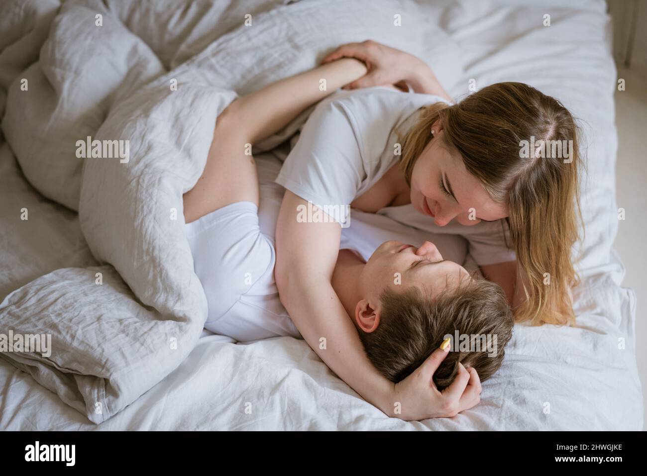 Happy young couple lie in bed and hug each other on white bed linen, look into each other's eyes
