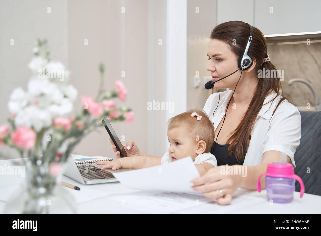 Mother with baby calling on smartphone while working at laptop Stock