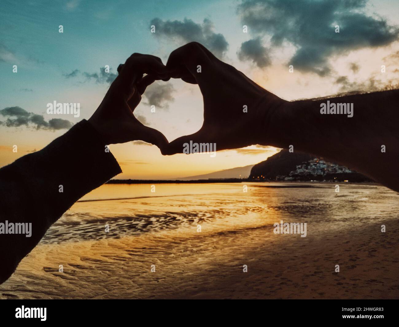Pov of couple doing heart gesture symbol with hands together at the beach in summer holiday vacation with amazing sky and colorful sunset in backgroun Stock Photo