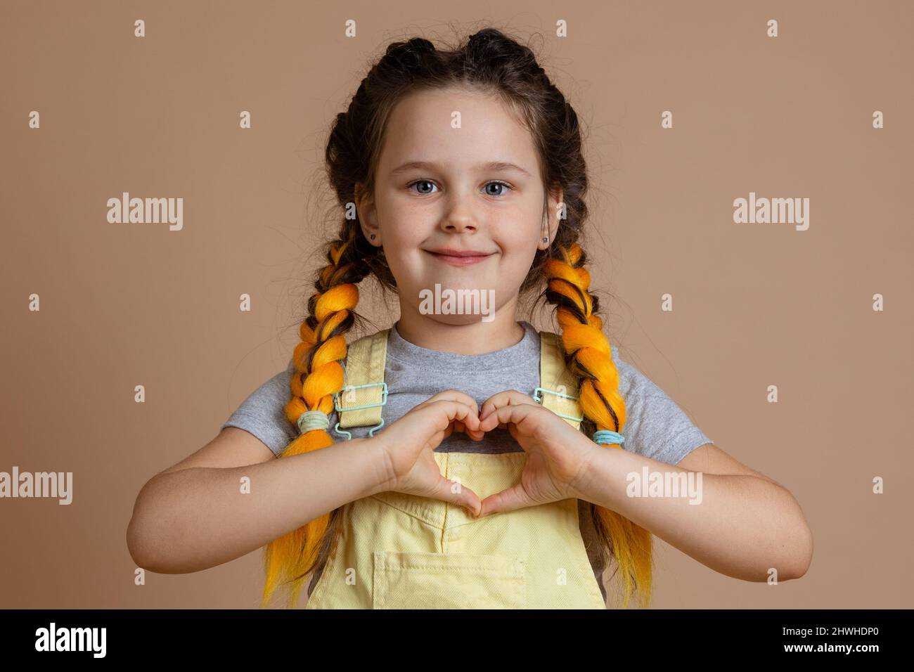 Kind little smiling girl, with yellow kanekalon pigtails looking at camera showing heart with fingers wearing yellow jumpsuit and gray t-shirt on Stock Photo