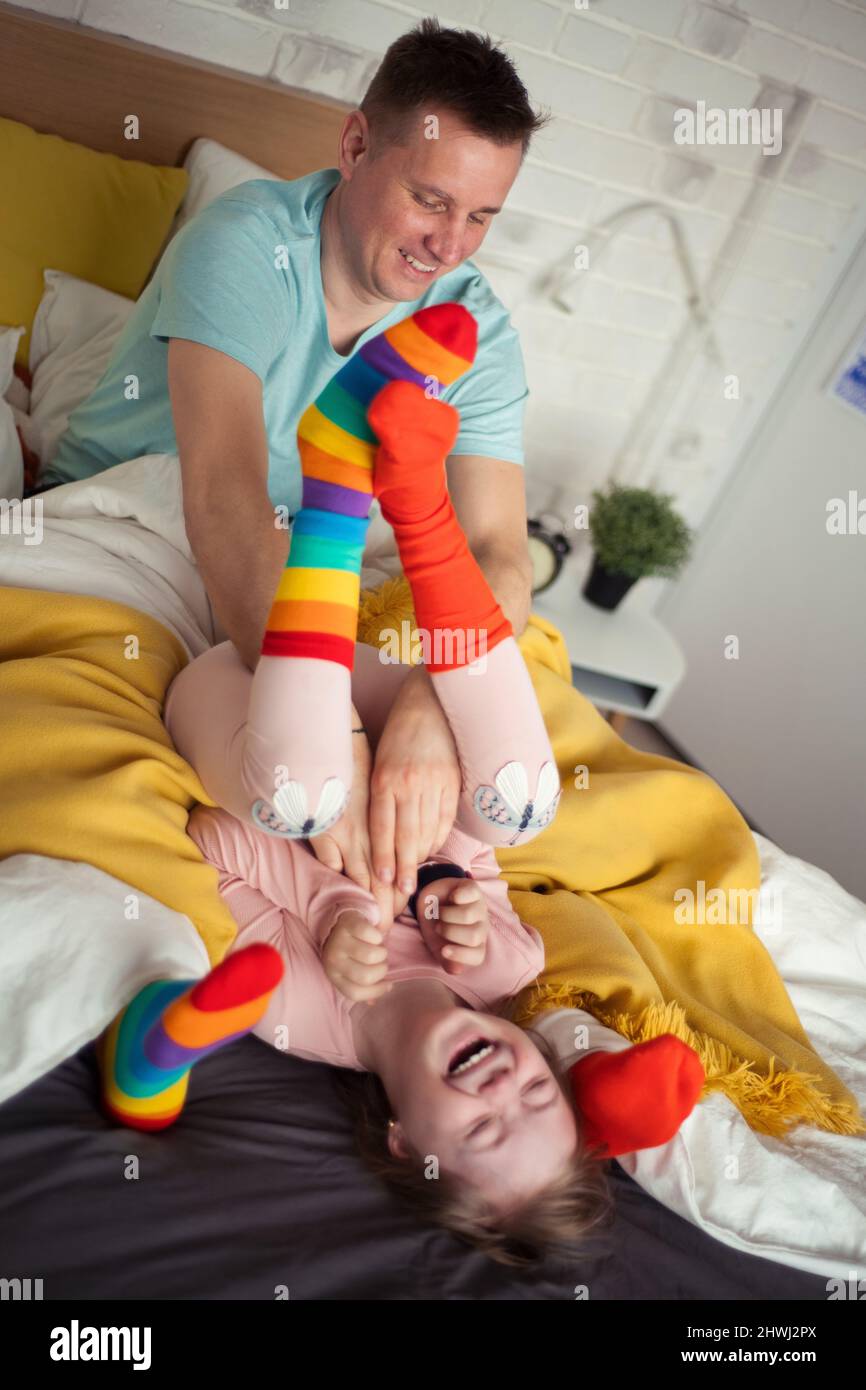 Cheerful little girl with Down syndrome lying on bed and laughing when her father is tickling her. Stock Photo