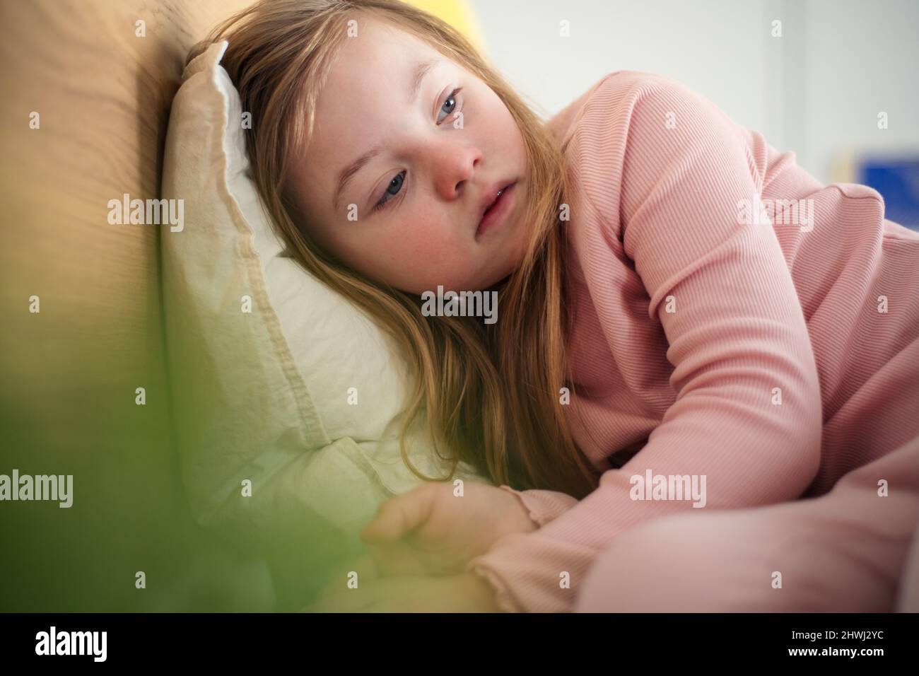 Sad little girl with Down syndrome lying on bed at home. Stock Photo