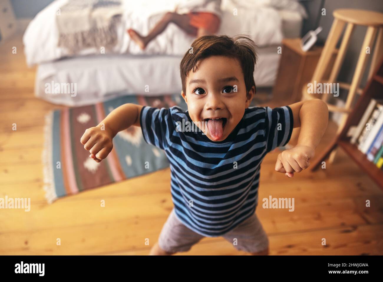 Cute young boy flexing his strength while standing in a bedroom with his sister in the background. Adorable little boy sticking his tongue out playful Stock Photo