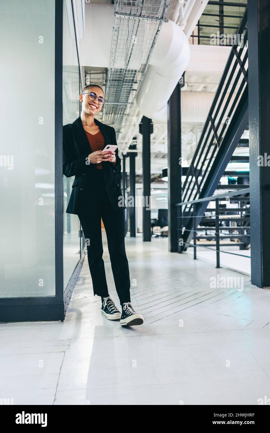 Cheerful businesswoman holding a smartphone in an office. Happy young businesswoman smiling at the camera while standing in a modern workplace. Young Stock Photo