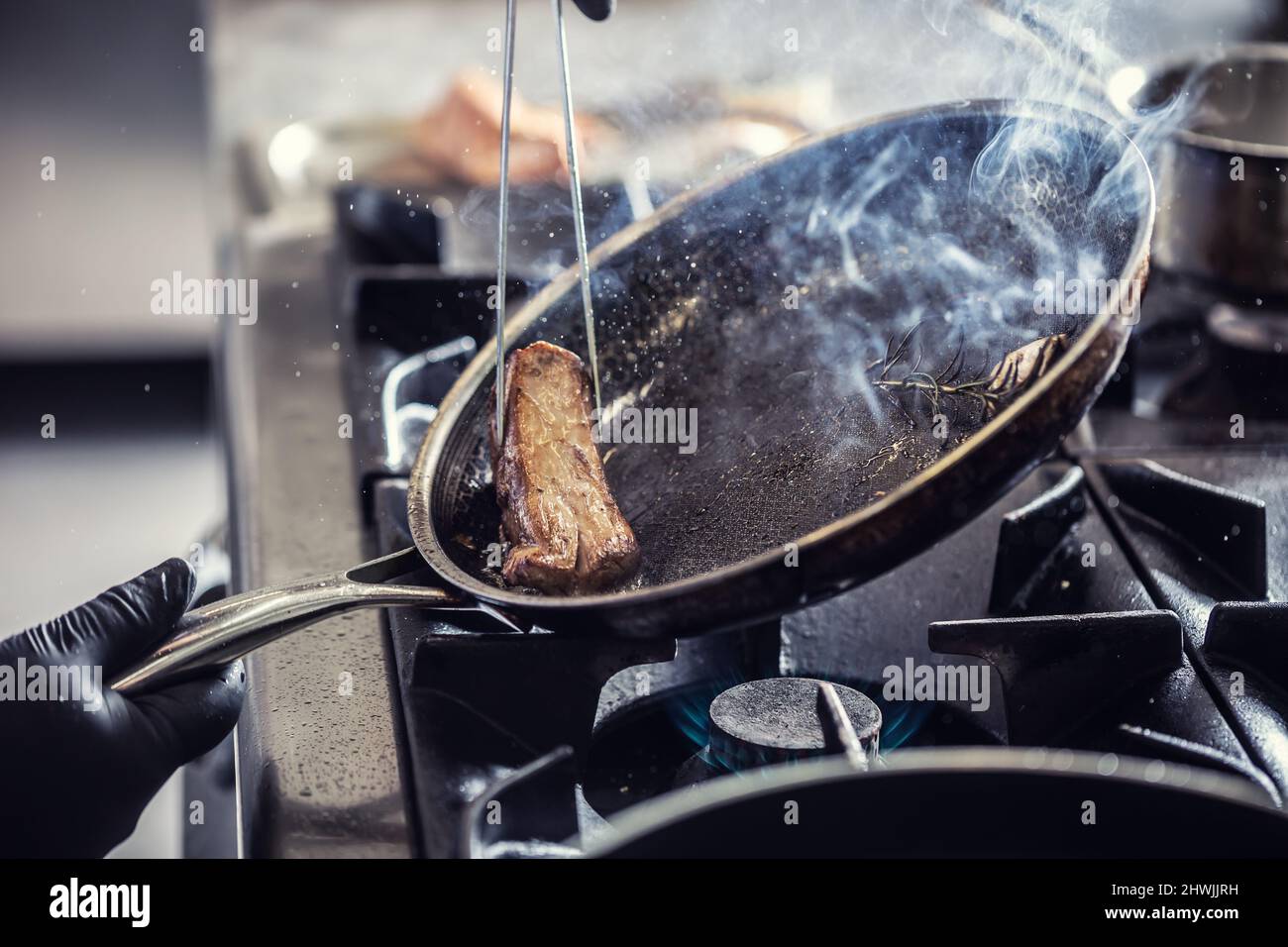 Detail of a pan with garlic and rosemary leaned to a side in order to pick a piece of meat with tweezers. Stock Photo