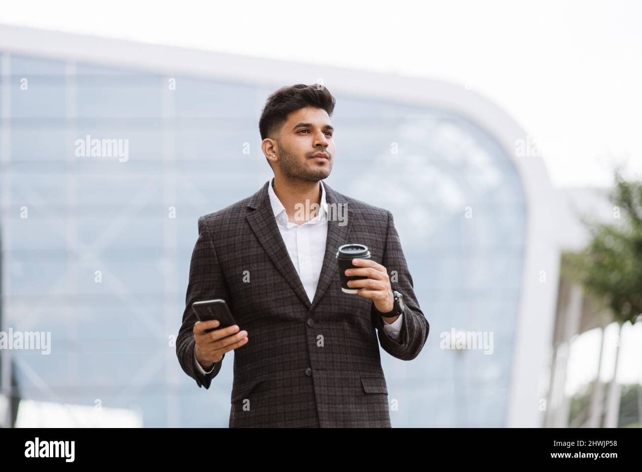 Handsome Indian businessman in formal wear, holding a cup of coffee looking away. Young thoughtful Arab man standing outside modern building and drinking take away coffee Stock Photo