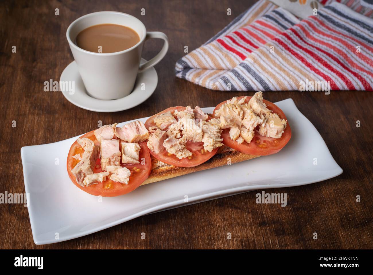 Toasted bread with sliced tomato and bonito tuna next to a cup of coffee with milk. Stock Photo