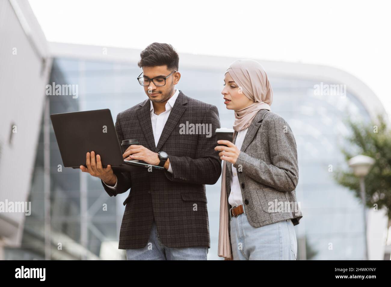 People and technologies concept. Attractive business couple of arab man and Muslim woman in smart casual style standing outside office working together on laptop smiling and discussing Stock Photo Ala...