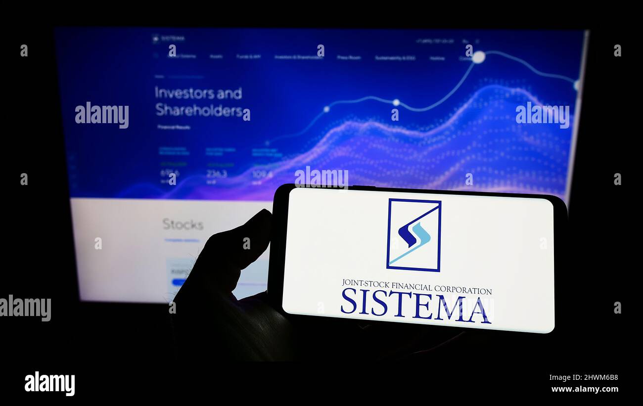 Person holding mobile phone with logo of Russian conglomerate AFK Sistema PAO on screen in front of business web page. Focus on phone display. Stock Photo