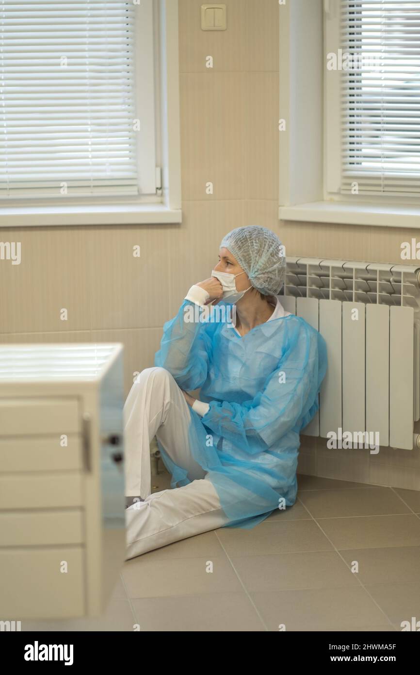 A tense female doctor sits tiredly on the hospital floor wearing a face mask and blue uniform. Stock Photo