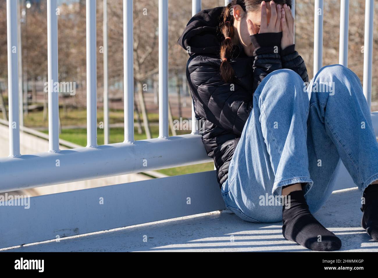 Girl hides her face with both hands sit on a bridge with white grids daytime Stock Photo