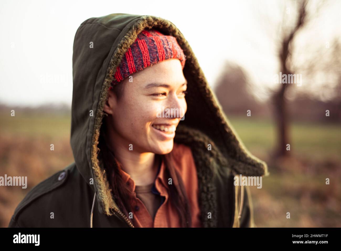 non-binary person in warm jacket hood smiles in afternoon sun outdoors Stock Photo
