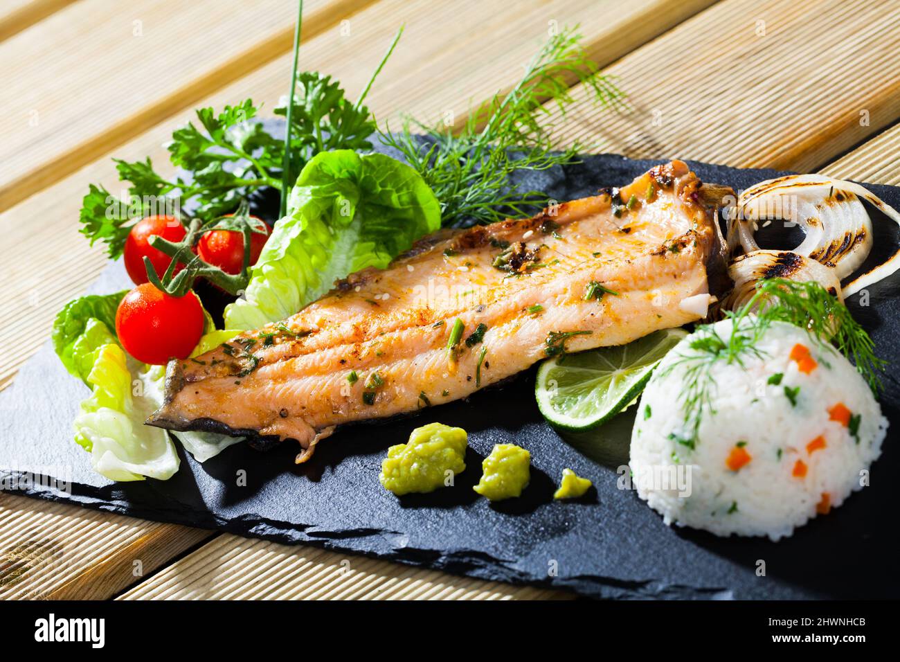Tasty fried trout fillet with rice, tomatoes and greens on black plate