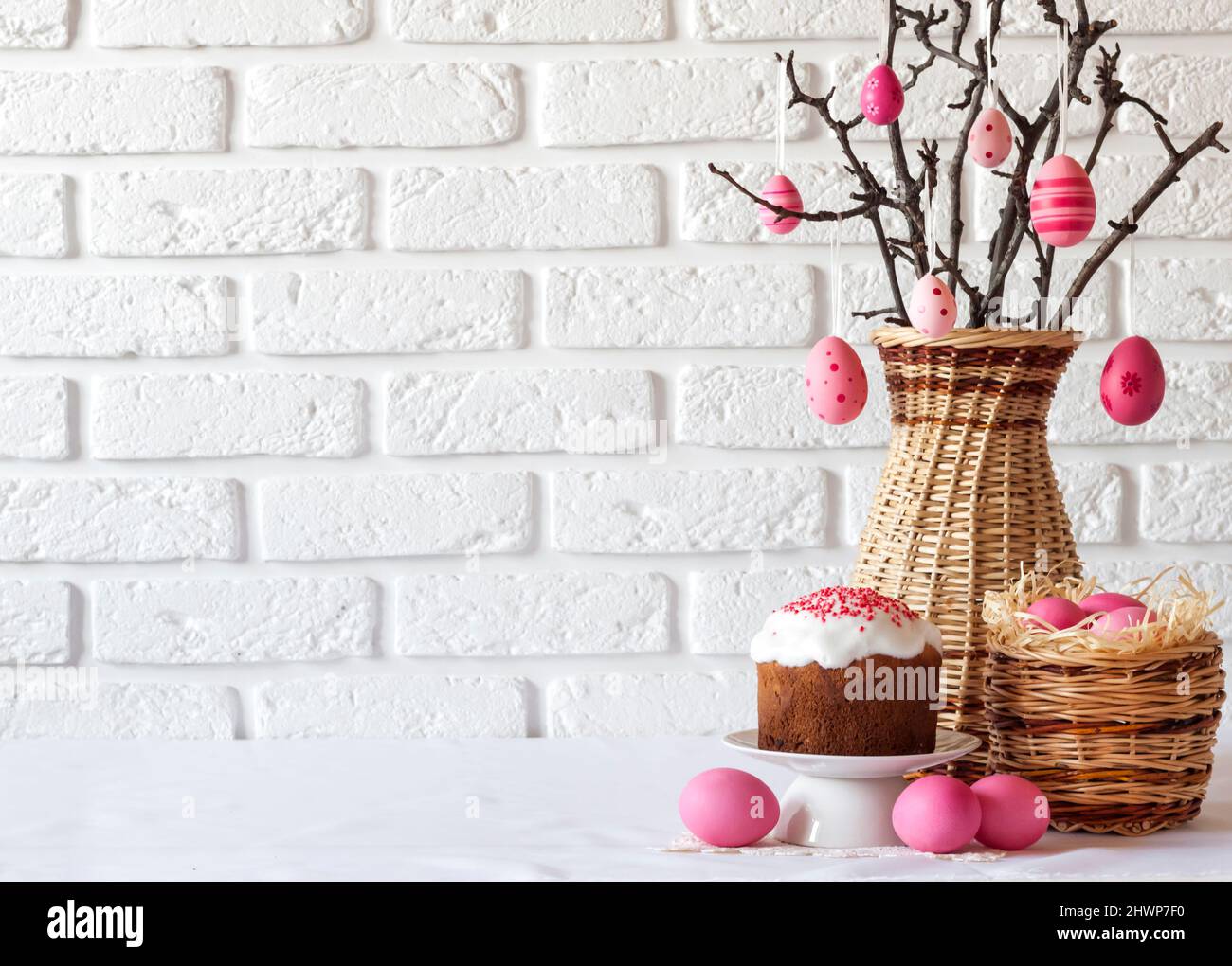 Easter composition with decorated tree branches in a wicker vase, pink colored eggs in wicker basket and Easter cake on white background. Copy space Stock Photo