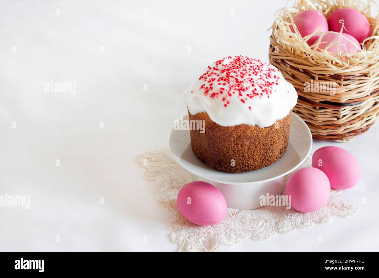 Minimalistic Easter composition with wicker basket with pink colored eggs and Easter cake on white background. Copy space Stock Photo
