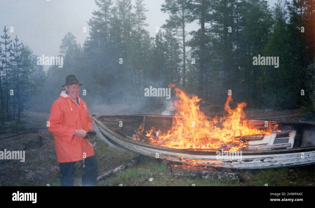 Smiling middle aged man standing next to old dilapidated burning rowing boat in forest, Sweden Stock Photo
