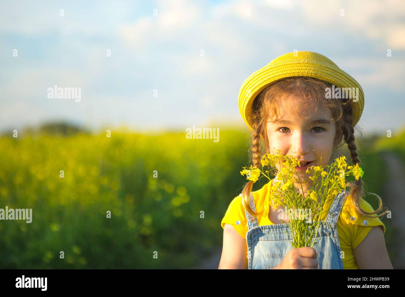 A girl in a yellow summer field sniffs a bouquet of flowers. Sunny day, holidays, allergy to flowering, freedom. Cultivation of agricultural rapeseed, Stock Photo