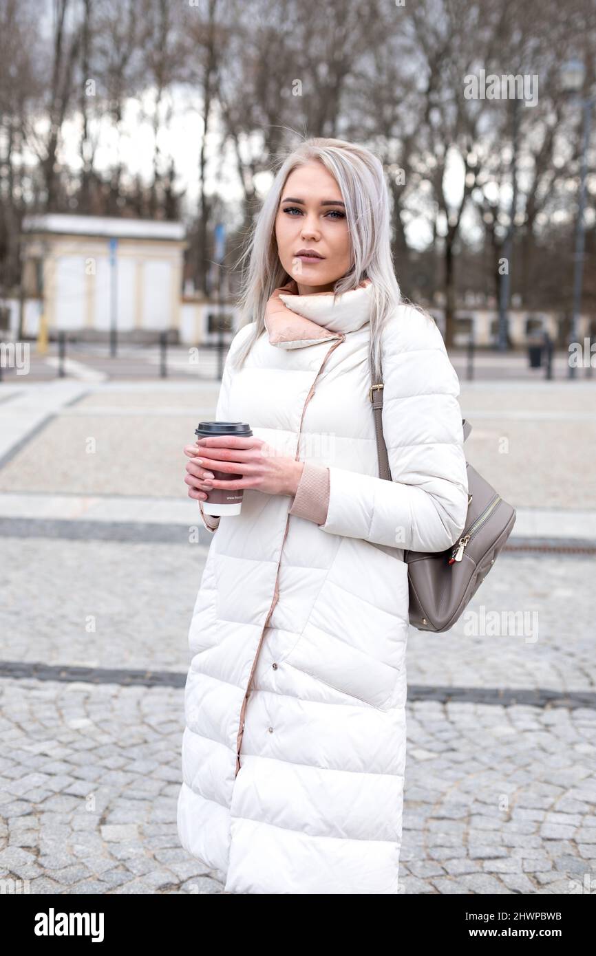 Vertical blonde attractive woman, staying look at camera, in warm white jacket, leather bag pack, drinking cup of coffee Stock Photo