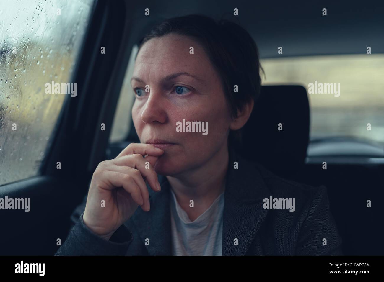 Disappointed and worried businesswoman looking out the window of taxi vehicle on cold rainy day on her way to work, selective focus Stock Photo