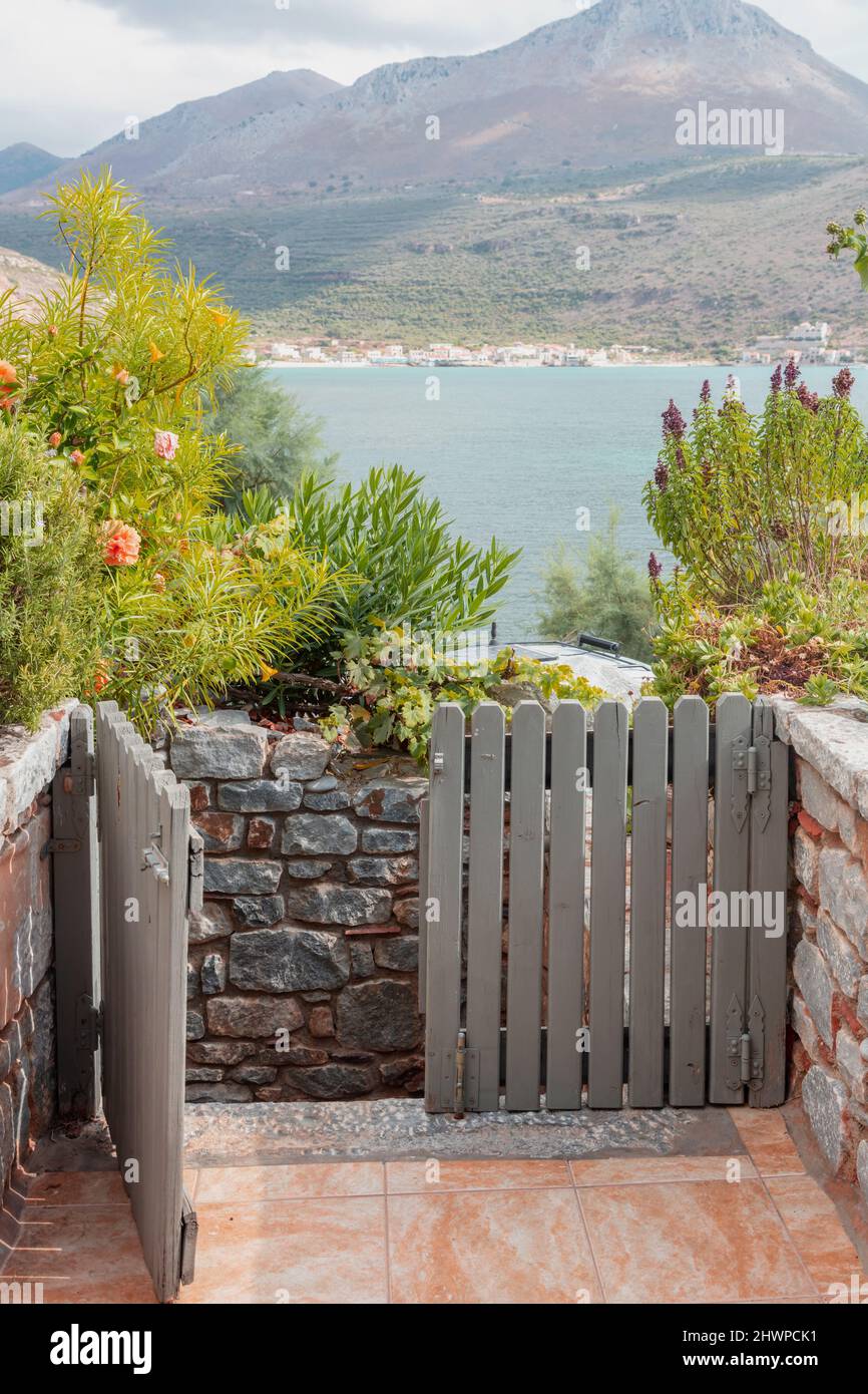 House grey fence gate open welcomes visitor. Calm Greek sea, mountain, building at seaside, blue sky view from home entrance over rocky wall through b Stock Photo