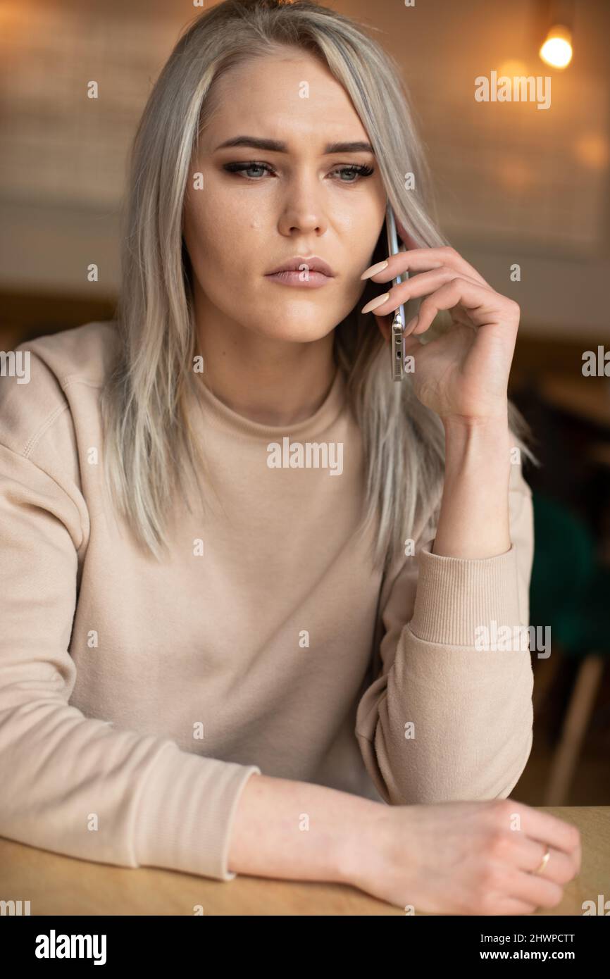 Vertical of dramatic surprised blond woman, staring, talking phone, cafe, sitting table. Business problem, remote work Stock Photo