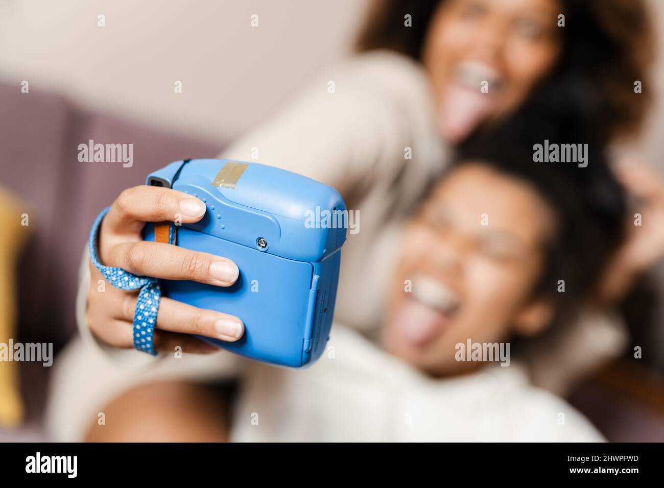 Two happy young african women friends taking selfies with an instant camera sitting on a couch at home close up grimacing Stock Photo