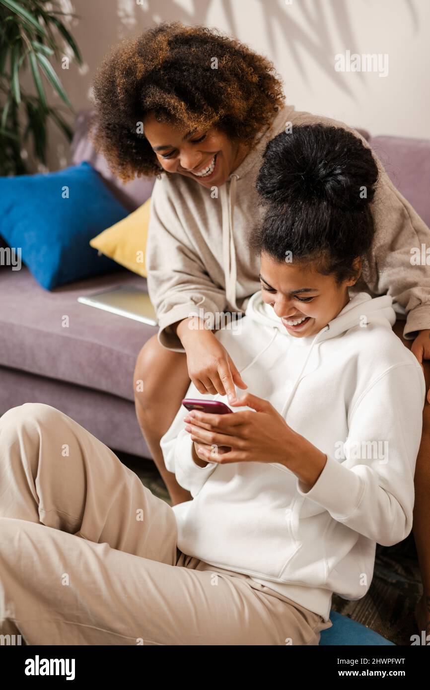 Two happy young african women sitting on a couch at home holding mobile ...