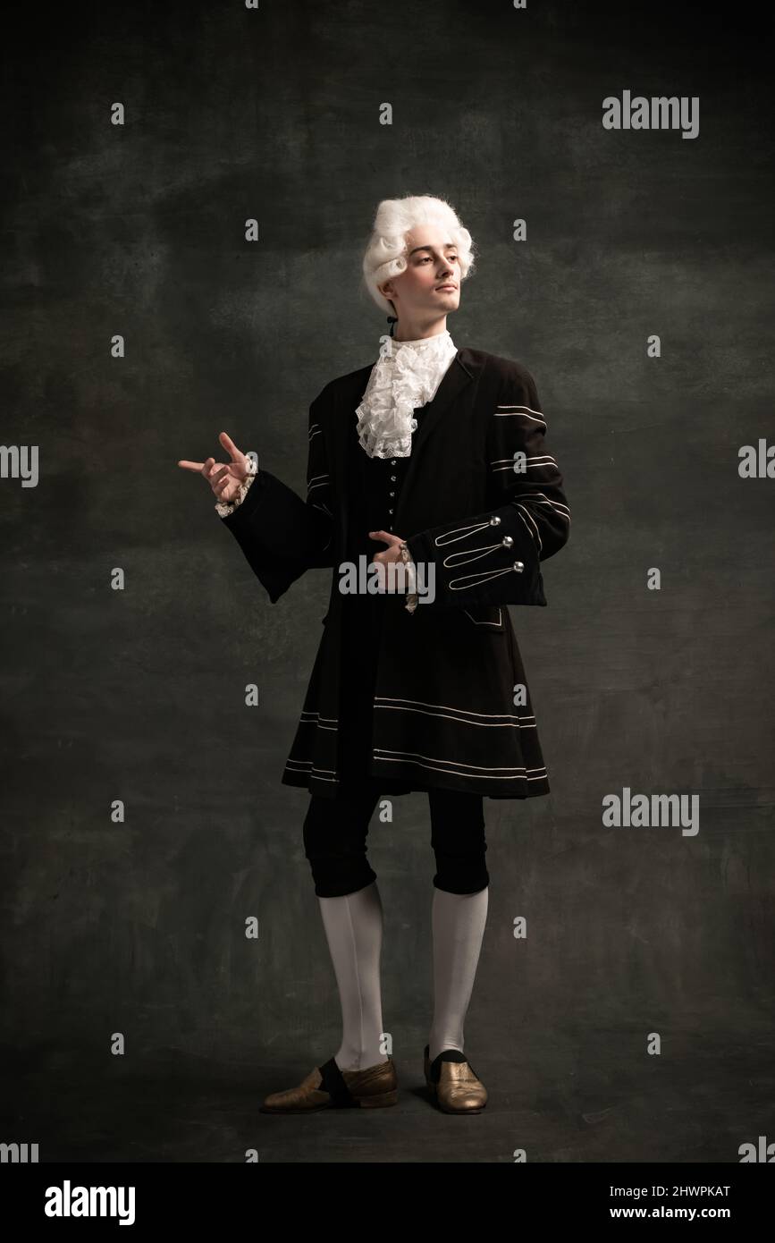 Young man wearing wig and vintage medieval outfit like famous composer isolated on dark green vintage background. Retro style, comparison of eras Stock Photo