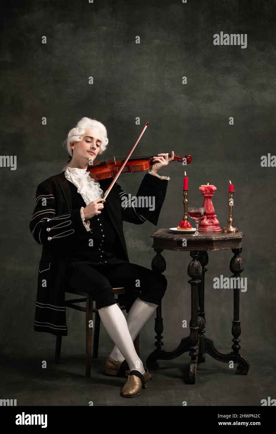 Portrait of young man wearing wig and vintage medieval outfit like famous composer playing violin isolated on dark green vintage background. Stock Photo