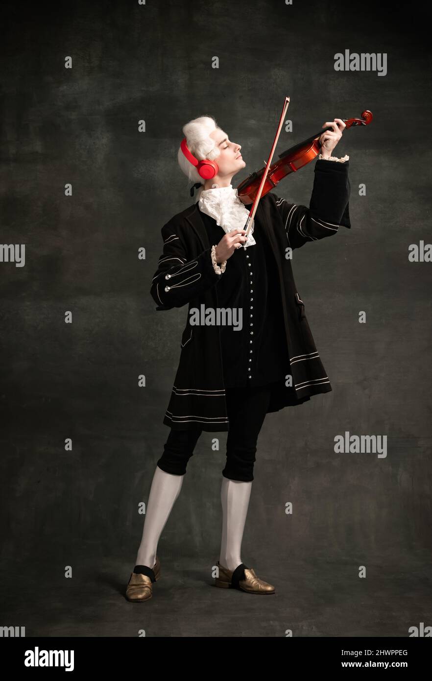 Portrait of young man wearing wig and vintage medieval outfit like famous composer playing violin isolated on dark green vintage background. Stock Photo
