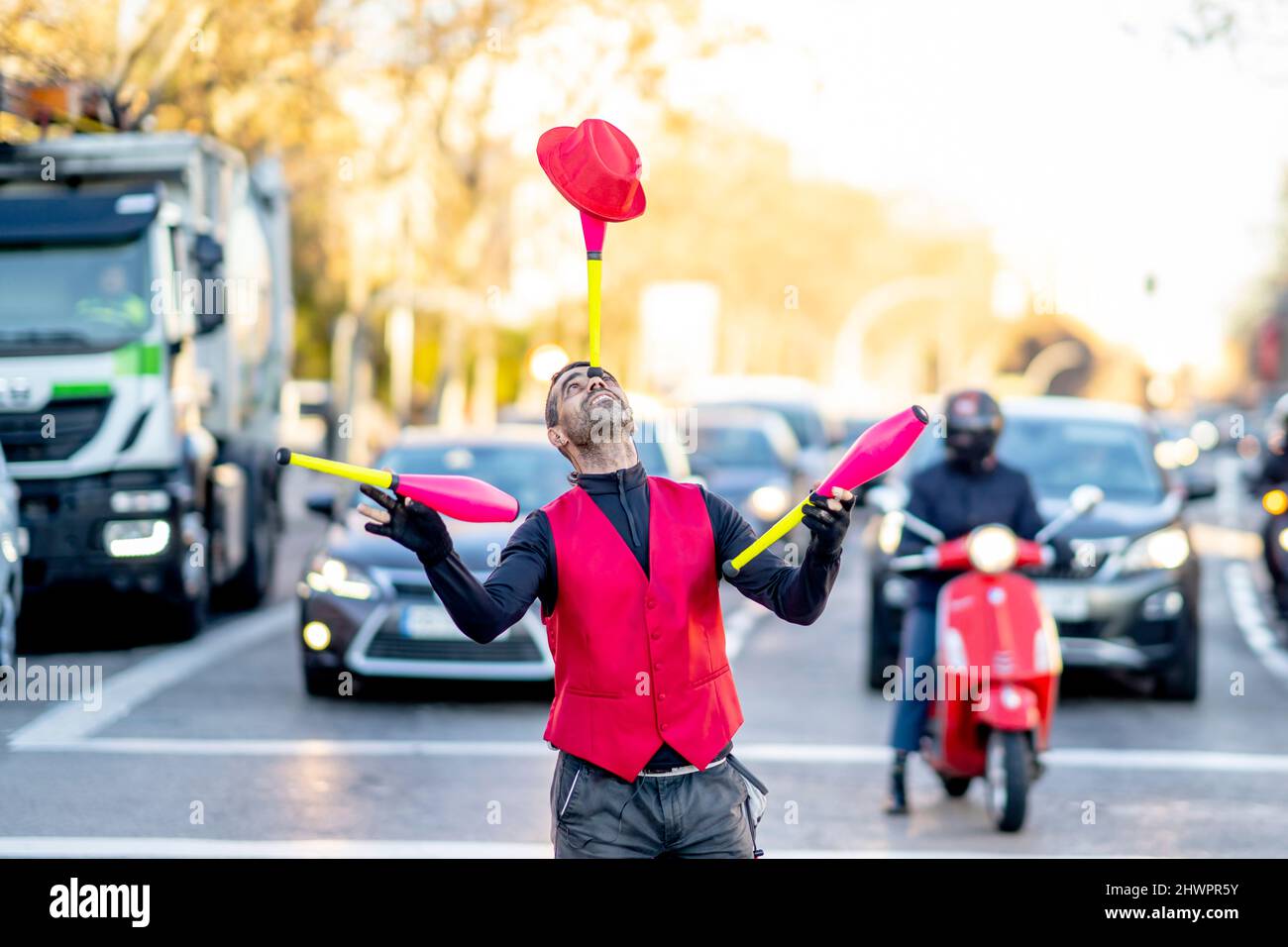 Performer balancing juggling pin and hat on nose in front of cars Stock Photo