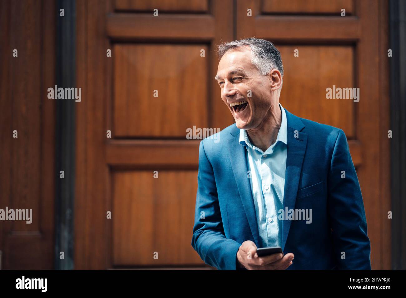 Businessman with smart phone laughing in front of closed door Stock Photo