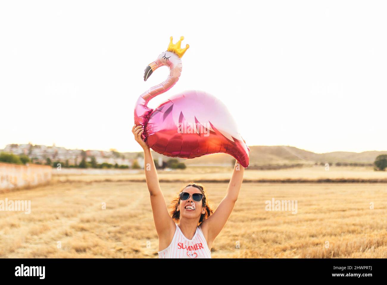 Smiling woman holding aloft flamingo balloon at sunset Stock Photo
