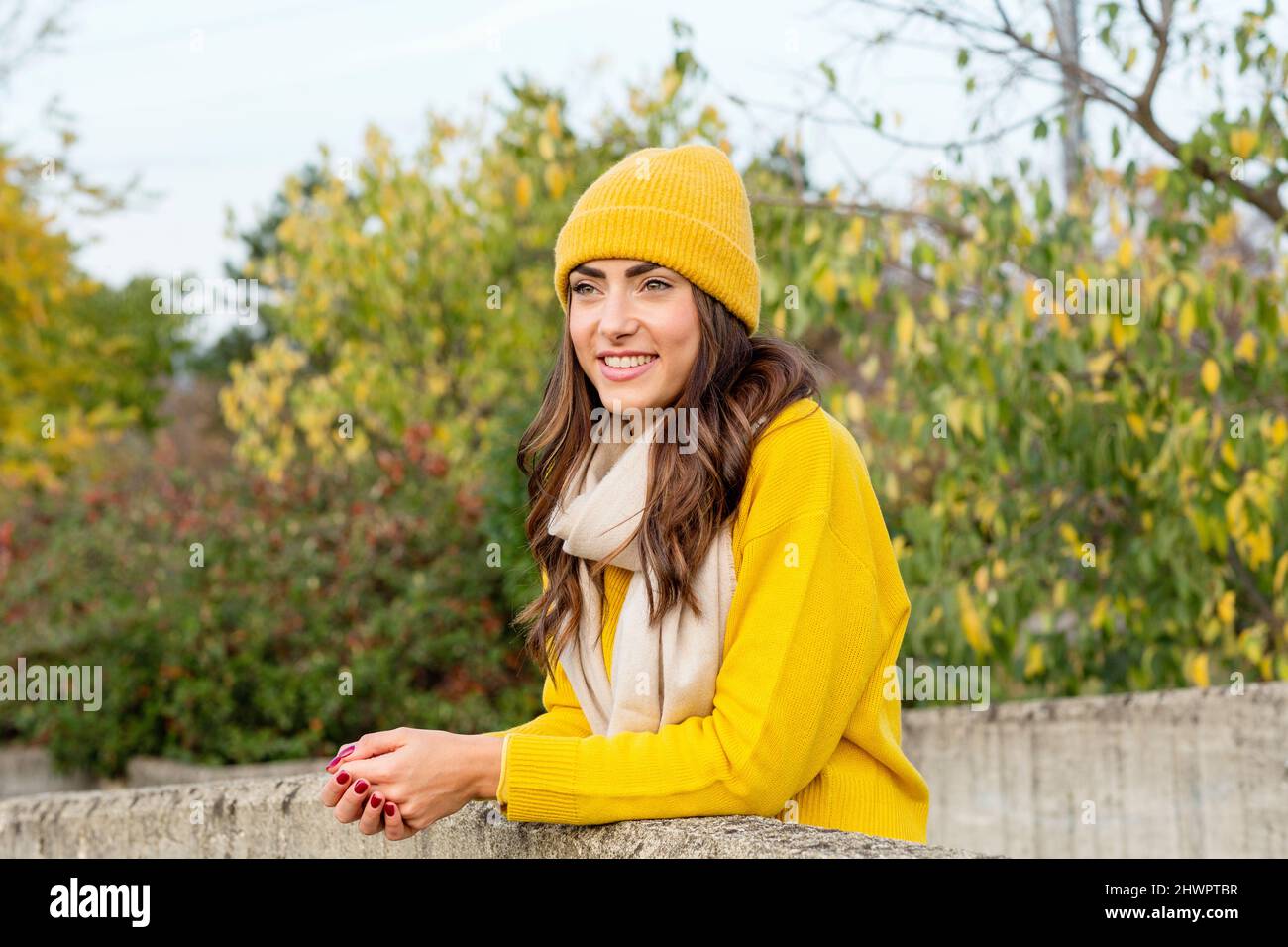 Young woman with yellow jumper and knit hat leaning on wall in park Stock Photo