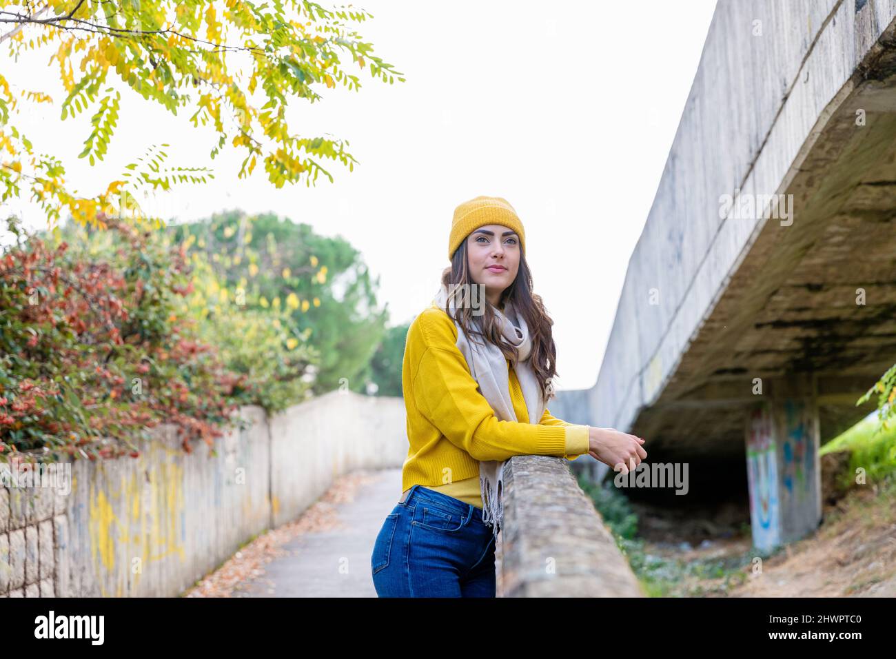 Young woman in yellow jumper and knit hat leaning on wall in autumn park Stock Photo